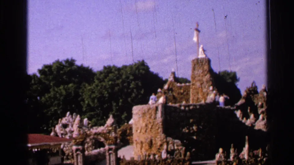 1965 Saint Louis 8mm frame showing visitors near an outdoor religious display with stone forms, statues, trees, and visible film wear.