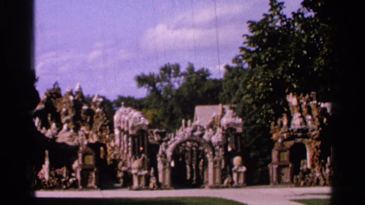 1965 Saint Louis frame showing arched stone niches, religious statues, a lawn, trees, and distant visitors at an outdoor devotional display.