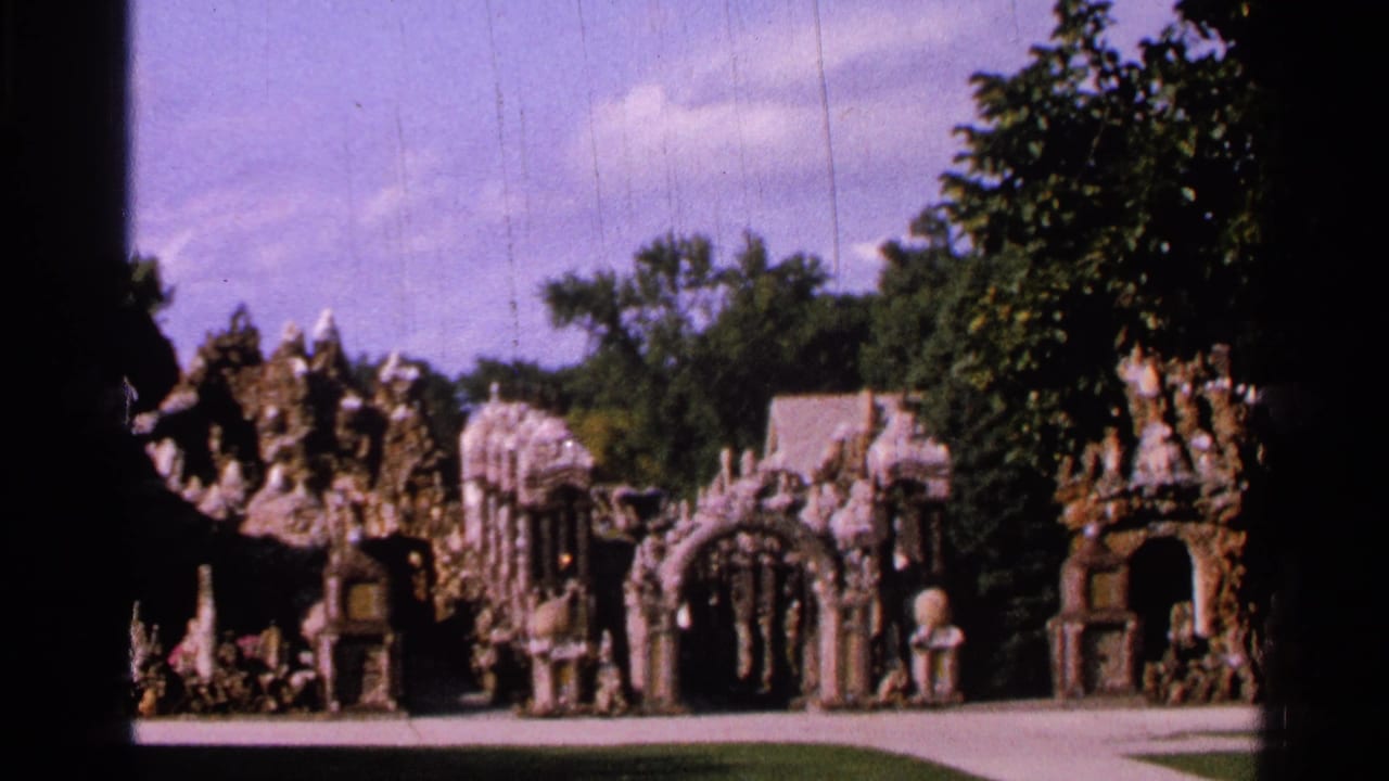 Wide view of ornate stone garden arches and tower-like structures beside trees and a paved path.