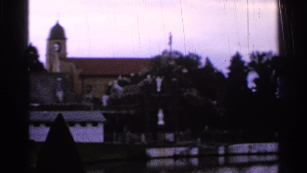 1965 Saint Louis 8mm frame showing a church steeple and roofline beyond trees, with dark foreground shapes and soft focus.