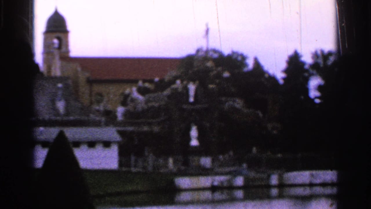 1965 8mm frame showing the church bell tower and red roof behind a grotto with white statues and trees.