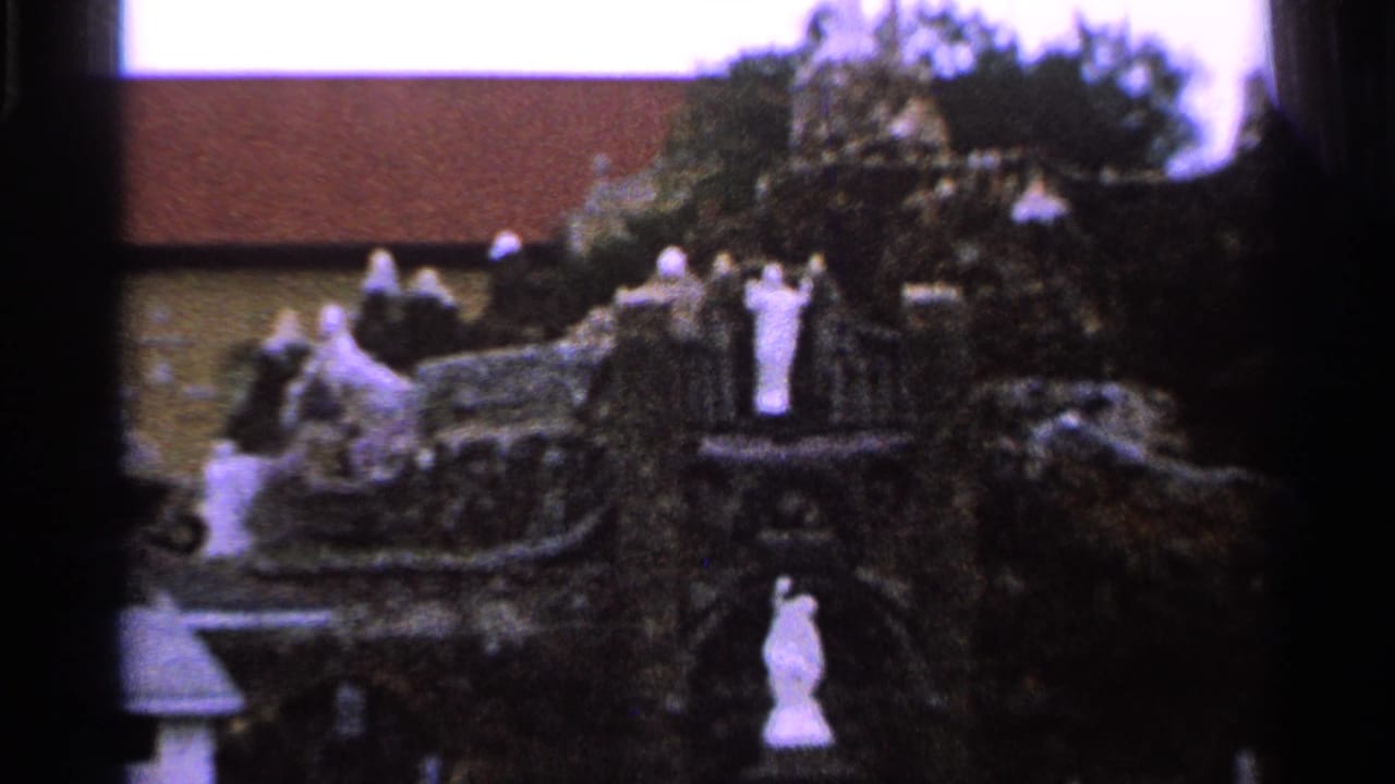 Close 1965 Saint Louis frame of a religious grotto with white statues, trees, and a church wall and roof behind it.