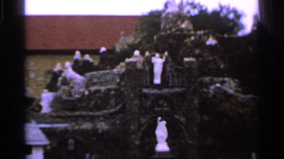Close 1965 Saint Louis frame of a religious grotto with white statues, trees, and a church wall and roof behind it.