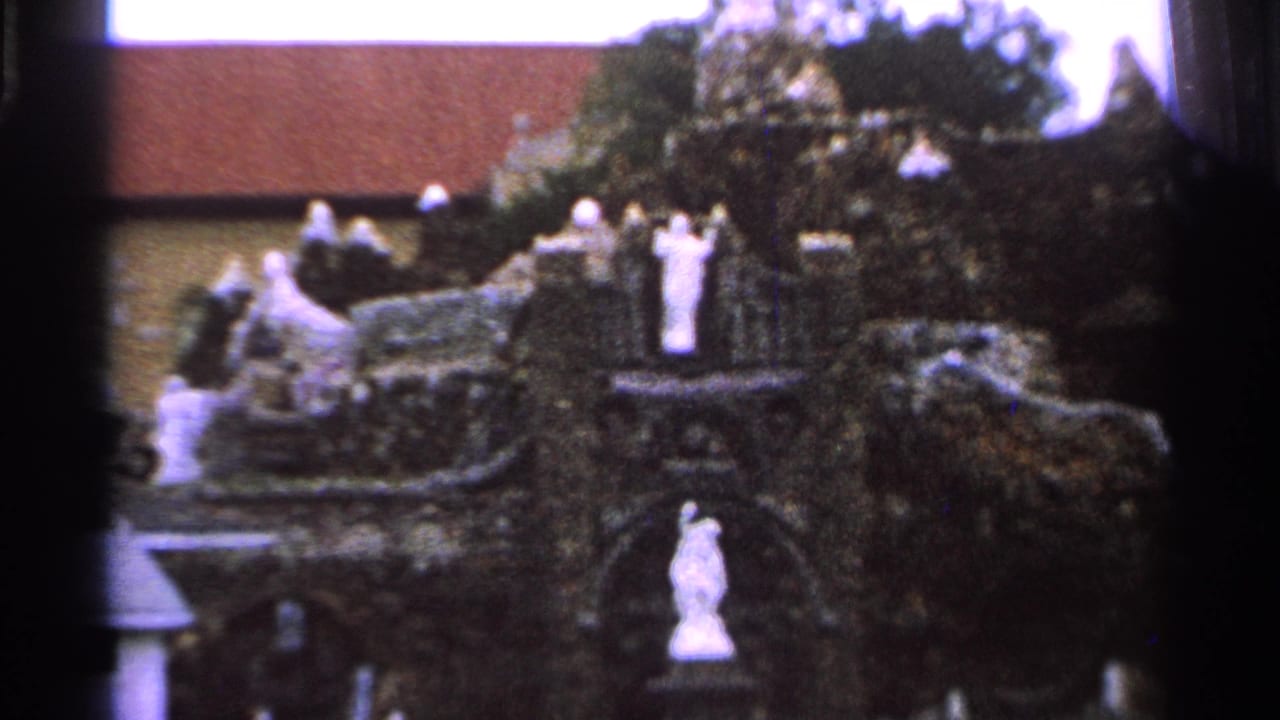 Decorative stone grotto with white statues, shrubs, and a red-roofed building behind it.