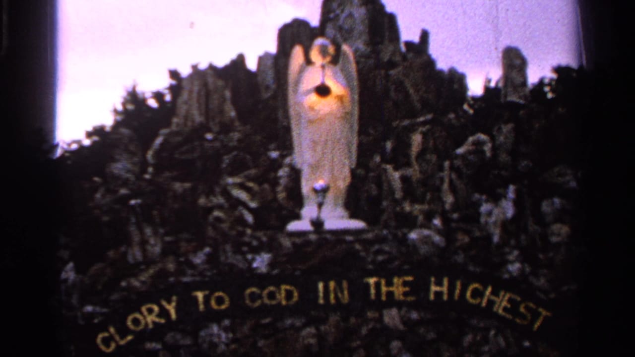 A devotional angel statue with a glowing heart stands above rocks and foliage, with GLORY TO GOD IN THE HIGHEST visible below.