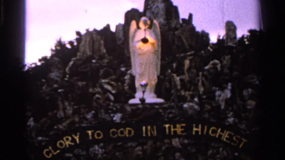 A devotional angel statue with a glowing heart stands above rocks and foliage, with GLORY TO GOD IN THE HIGHEST visible below.