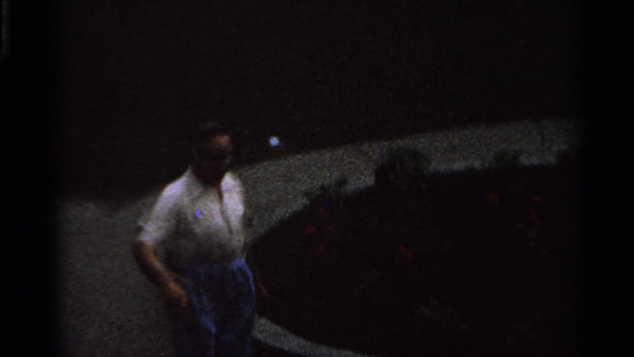 A dim 1965 Saint Louis 8mm frame shows an adult man walking along a curved path near flower beds and garden rockwork.