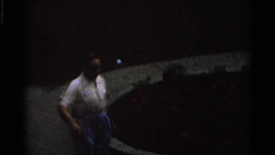 A dim 1965 Saint Louis 8mm frame shows an adult man walking along a curved path near flower beds and garden rockwork.