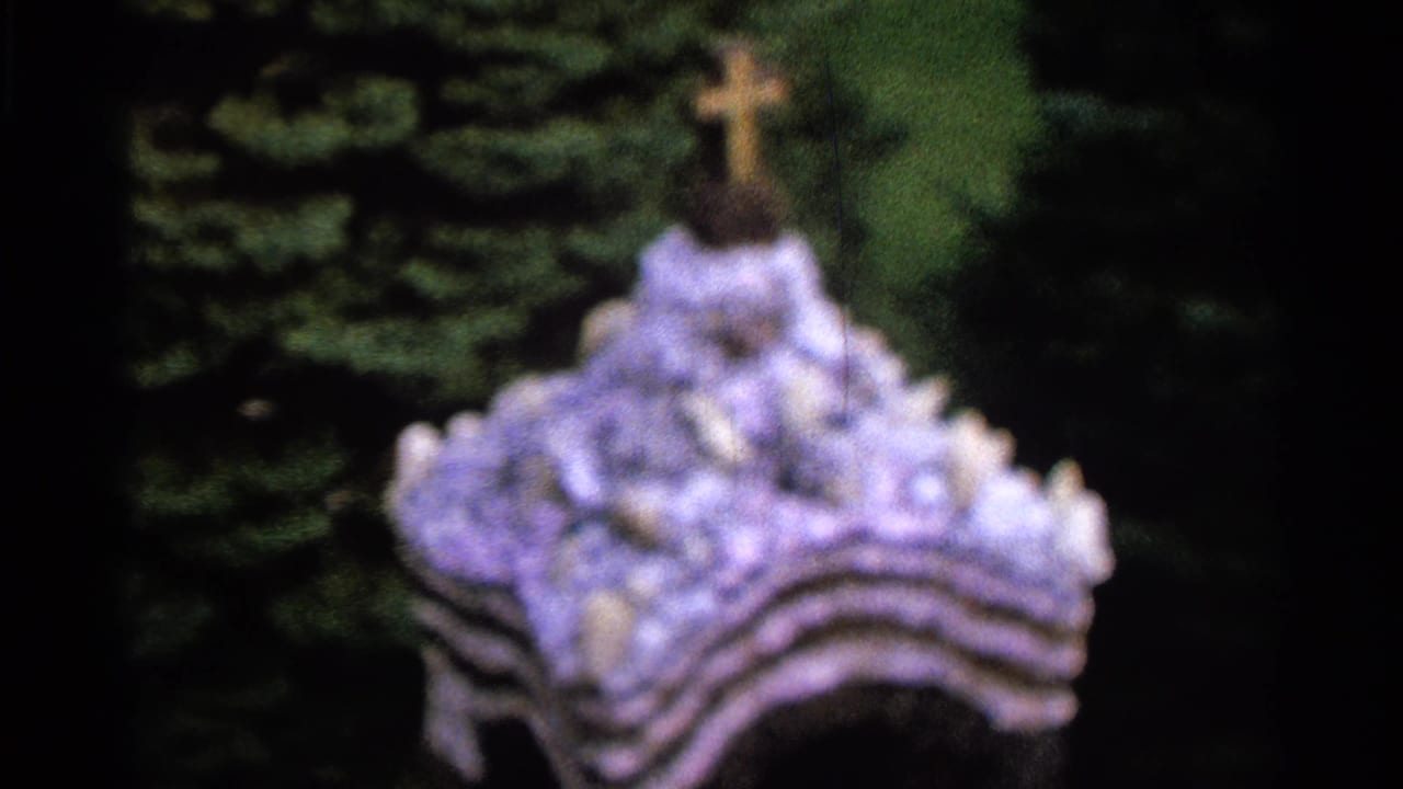 The final frame holds on the shrine's lower structure, showing the cross, pale decorative forms, greenery, and dark 8mm frame edges.