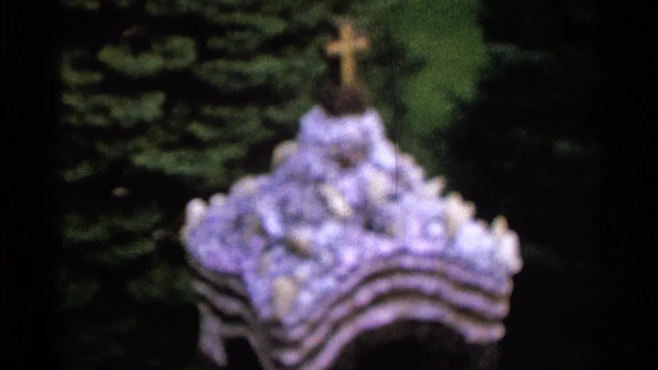 The final frame holds on the shrine's lower structure, showing the cross, pale decorative forms, greenery, and dark 8mm frame edges.