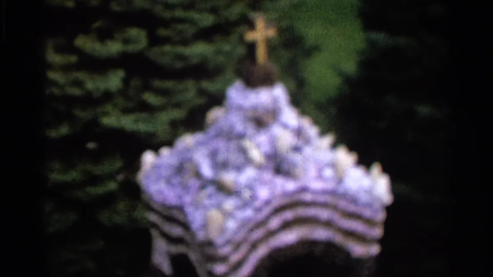 Soft 1965 Saint Louis 8mm frame showing a flower-covered cross or shrine form with dark trees behind it.