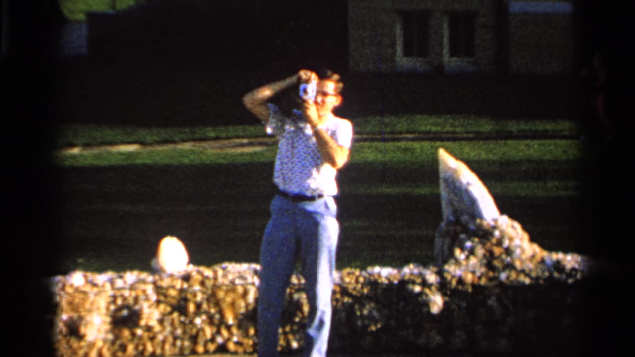 Adult man outdoors holding a camera near a low stone wall and lawn.