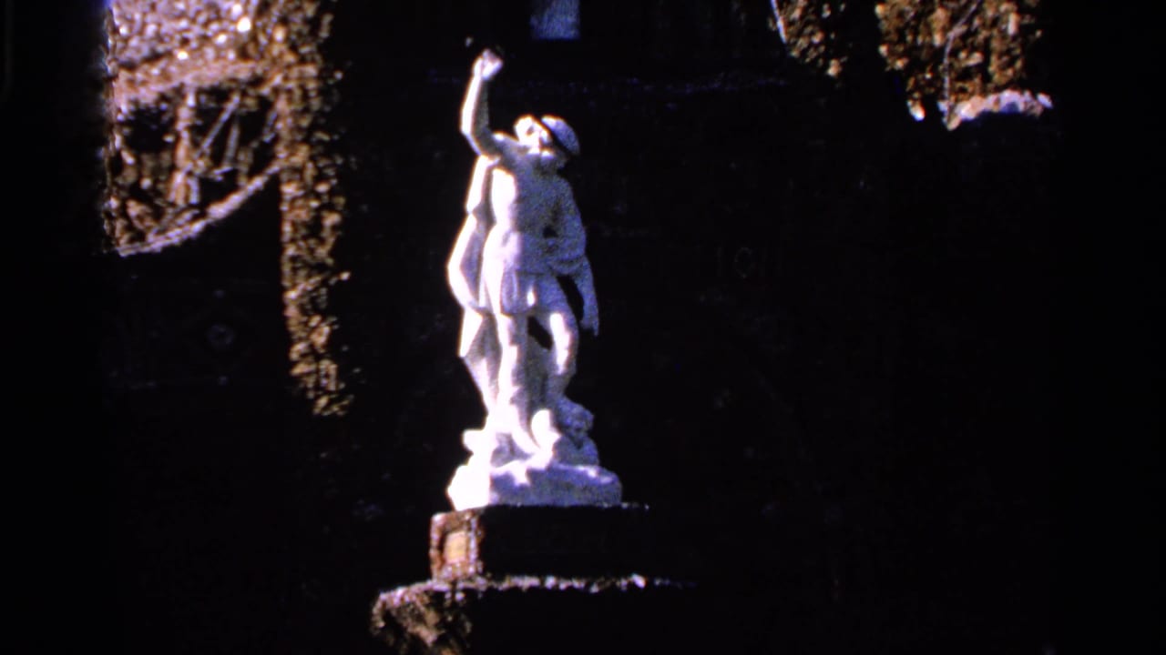 White statue on a pedestal against dark ornamental stonework and foliage.