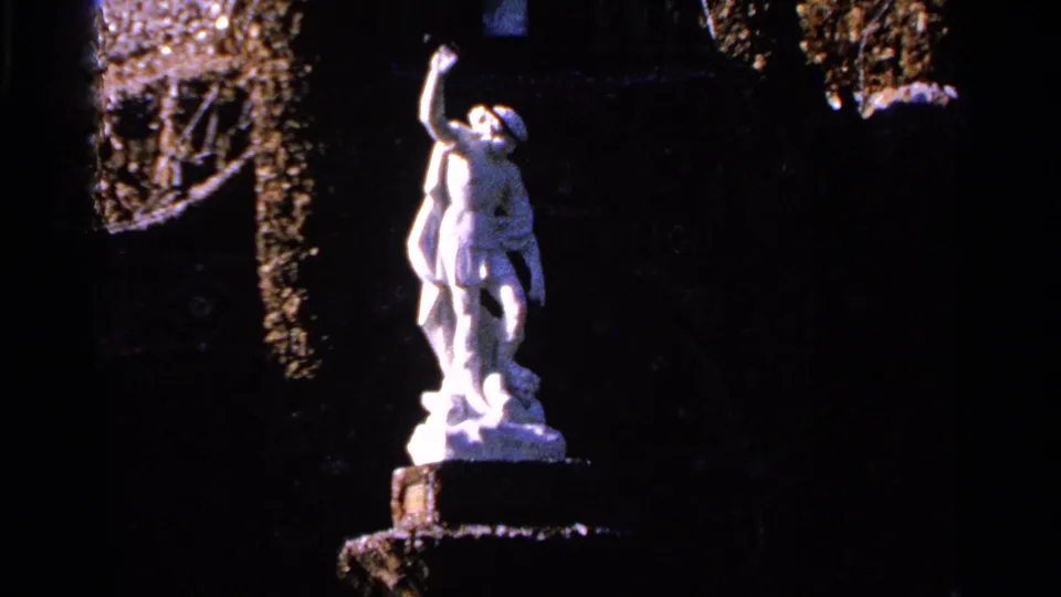 1965 Saint Louis home-movie frame showing a white classical-style statue on a pedestal in a shadowed landscaped setting.