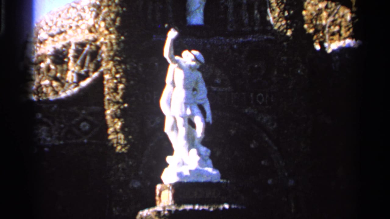 Close visitor-shot view of the white garden statue and pedestal, with ornate dark stonework and foliage visible behind it.