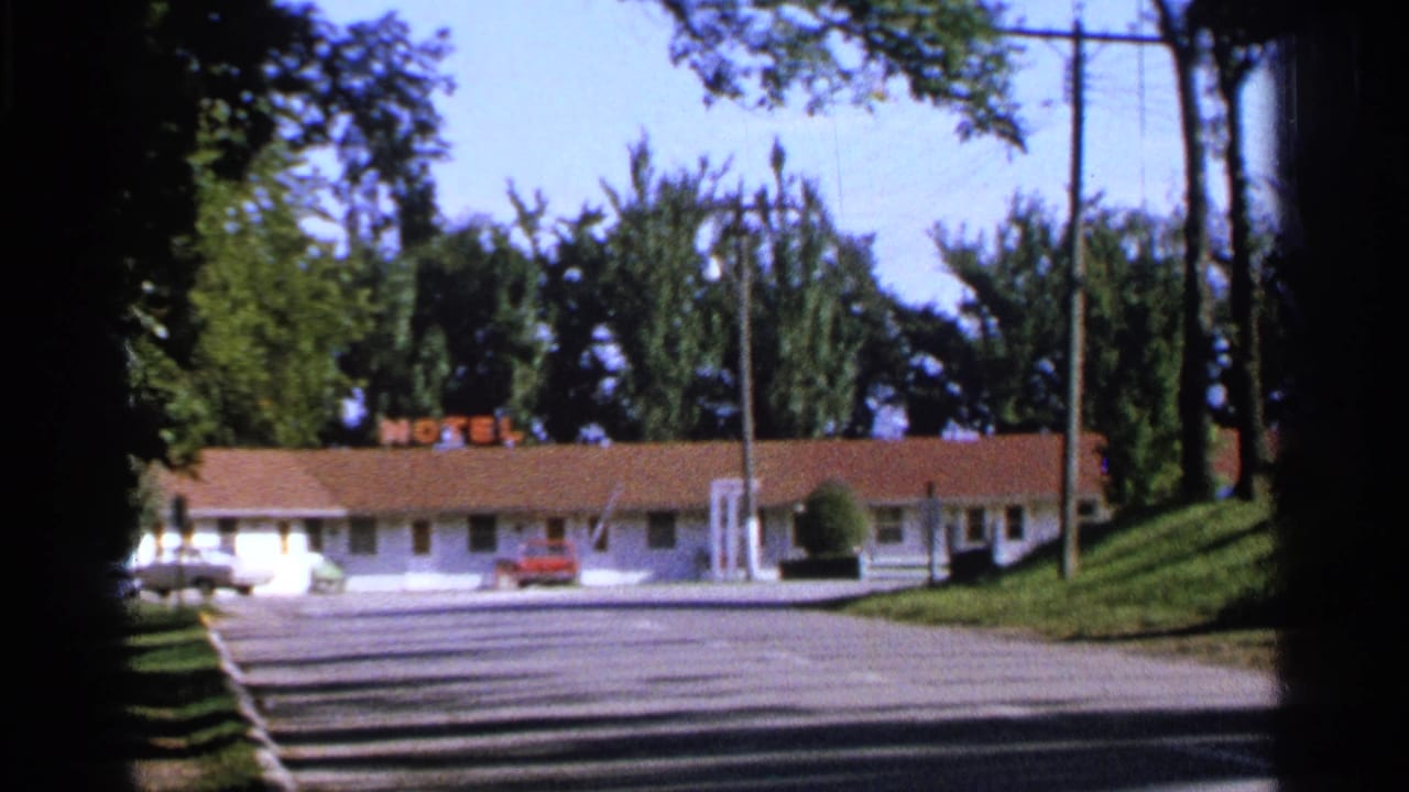 1965 Saint Louis frame showing a low roadside motel building beyond a road, with parked cars, trees, utility poles, and a motel sign.