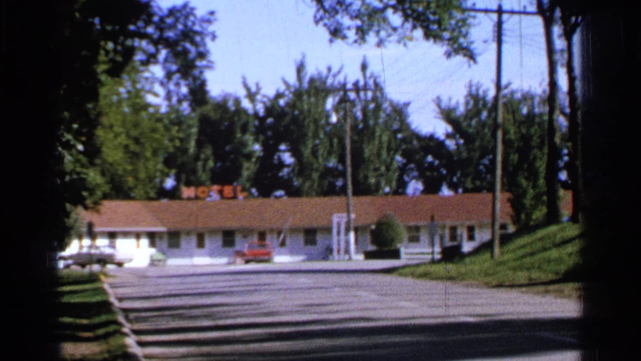 White roadside motel with red roof, neon MOTEL sign, parking lot, trees, utility poles, and parked cars.
