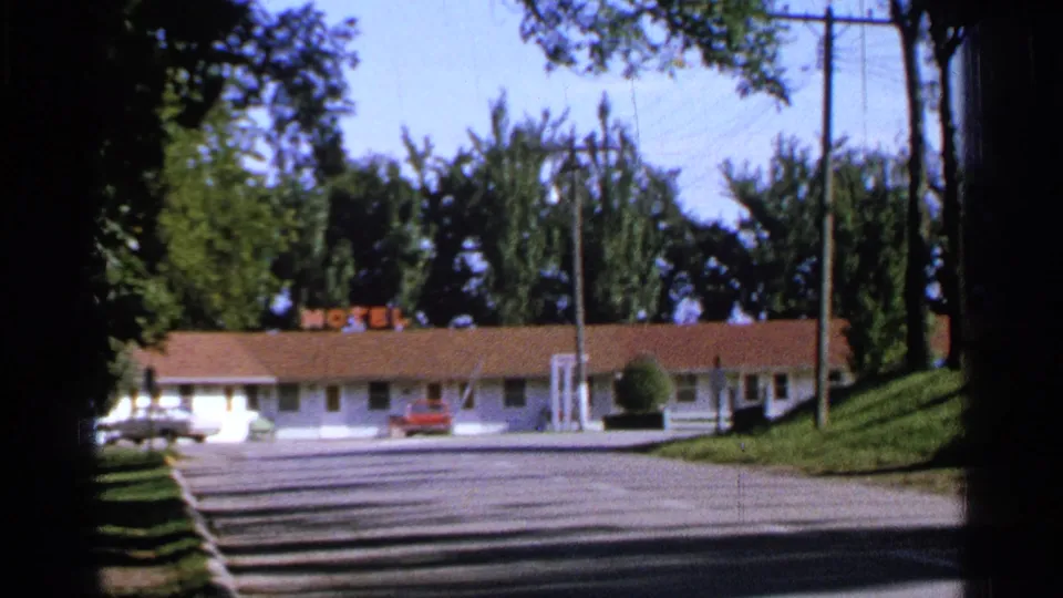 1965 home-movie view in Saint Louis showing a low white motel, red roof, neon MOTEL sign, empty parking spaces, trees, and classic cars.