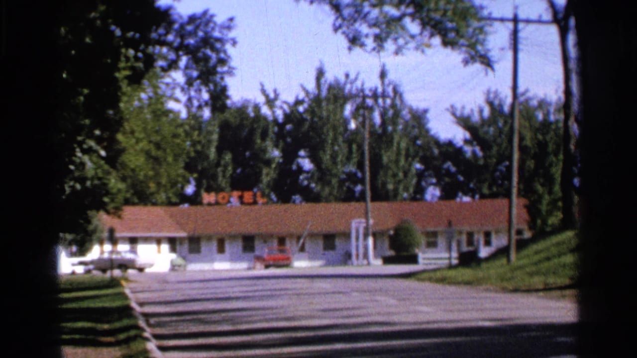 Roadside motel frontage with neon MOTEL sign, white building, red roof, parking lot, trees, and utility poles.