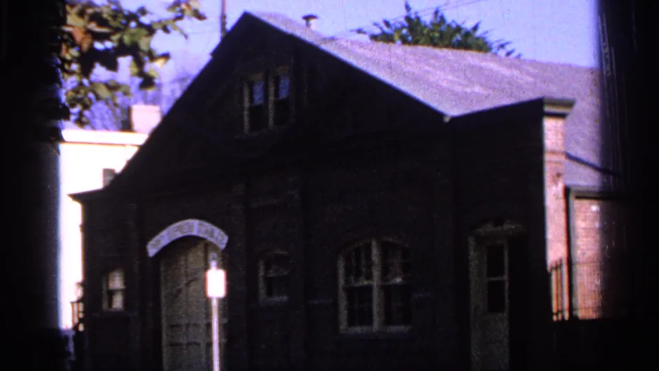 Saint Louis 1965 home-movie frame showing a dark brick building with an arched entry, windows, and neighboring structures nearby.