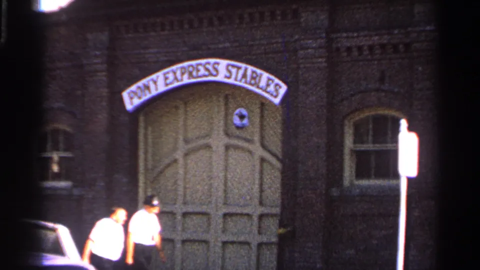 1965 Saint Louis home-movie frame showing two adult men in light shirts walking near the Pony Express Stables arched entrance.
