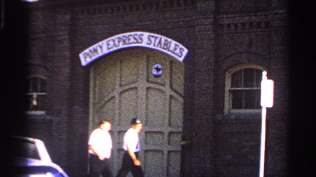Two adult men walk past a brick building entrance marked Pony Express Stables.