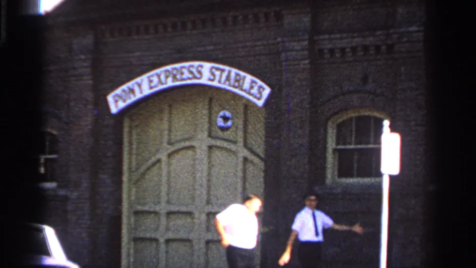 1965 Saint Louis street-level frame with two men beside the Pony Express Stables doors, brick wall, arched windows, and a signpost.