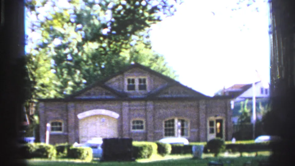 1965 Saint Louis exterior frame showing a brick building with garage-style doors, trees, hedges, parked cars, and strong daylight exposure.