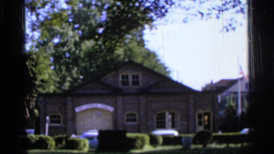 1965 Saint Louis 8mm frame of a brick garage-front building partly shaded by trees, with hedges and parked cars along the frontage.