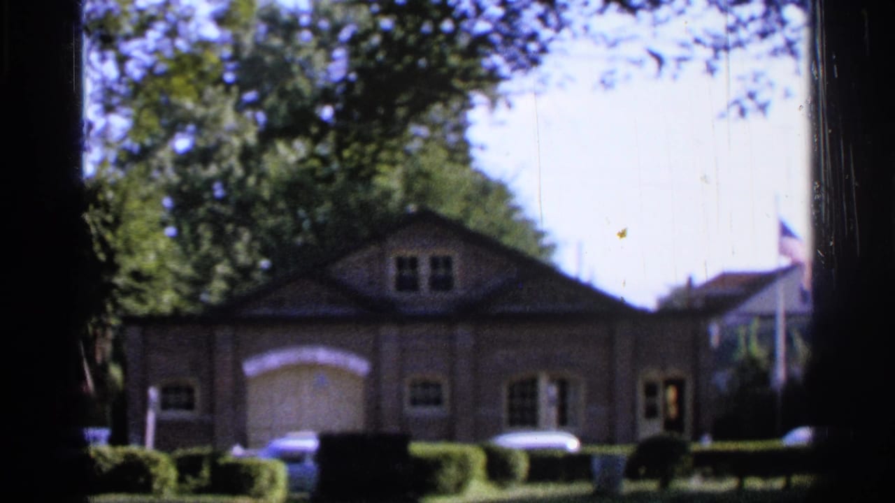 Front view of a brick house with trees, shrubs, and parked cars.