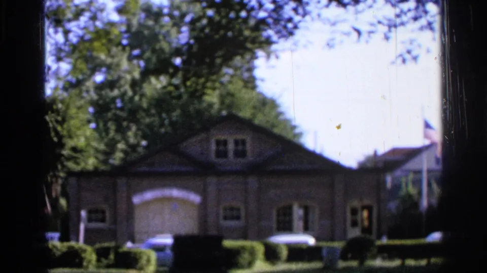 1965 Saint Louis home-movie frame showing a brick house from the front, framed by trees, shrubs, and parked cars.