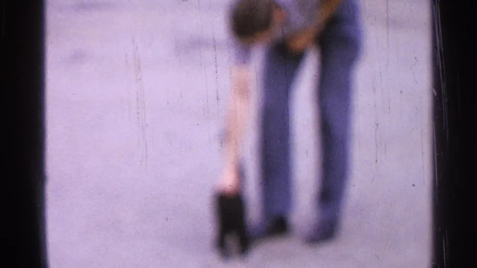 Soft-focus 1965 home-movie frame of a man in casual clothes bending toward a small black cat on a paved surface.
