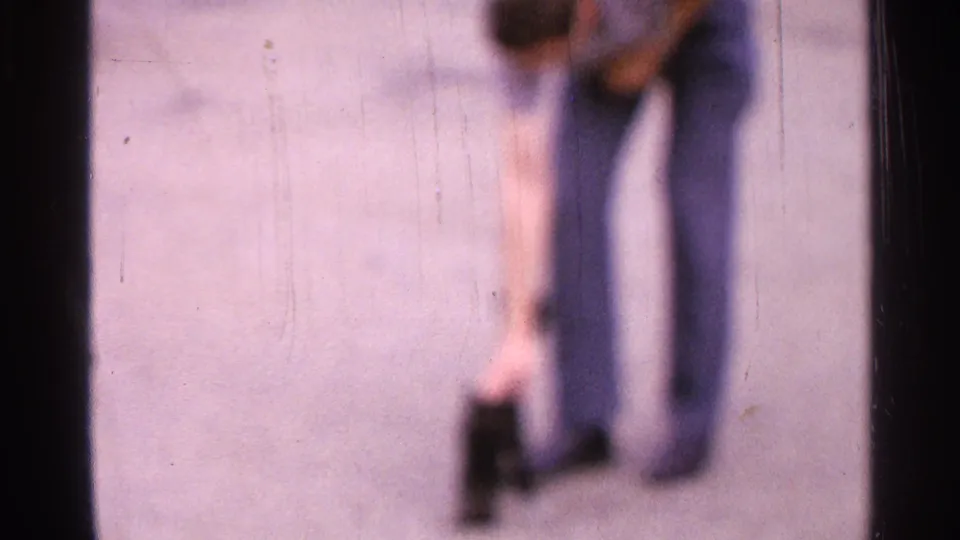 Blurred 8mm frame from Saint Louis showing a casually dressed man reaching down near a small black cat on pavement.