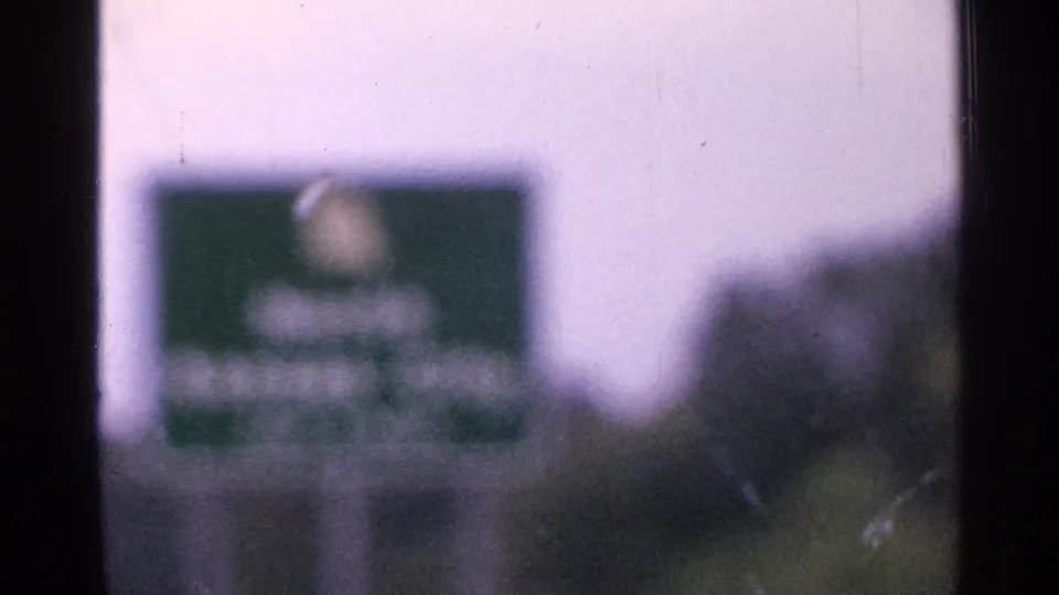 Close, unreadable green road sign in a 1965 Saint Louis driving shot, softened by motion and 8mm grain.