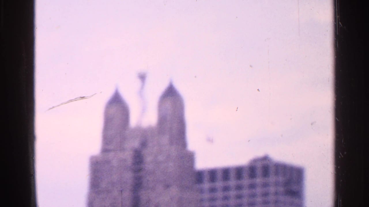 Soft 1965 8mm frame showing a distant twin-spired Missouri landmark with pale sky and visible film edges.
