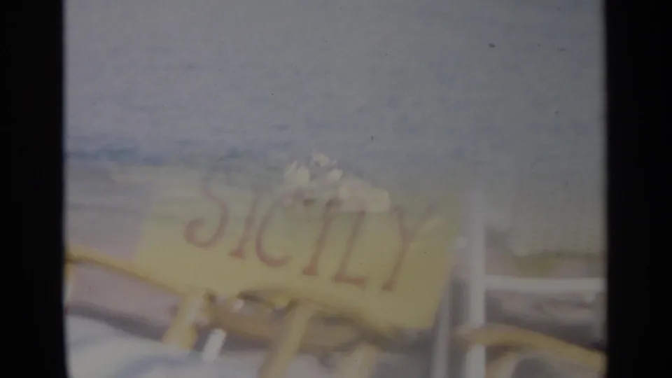 Partially overlaid 8mm frame where the sea view fades into a yellow hand-lettered Sicily sign near beach seating.