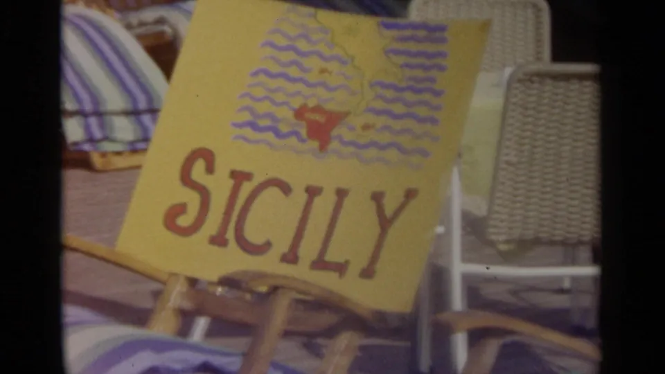 Yellow hand-drawn Sicily sign in red lettering, propped against beach seating with striped fabric nearby.