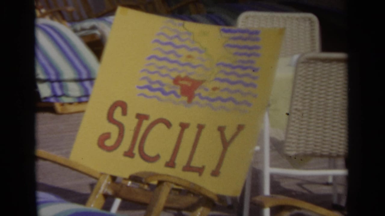 Yellow hand-drawn Sicily sign beside wicker chairs and striped towels.