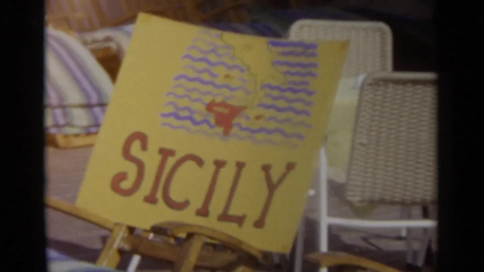 The Sicily sign remains in view, tilted on a wooden stand with wicker chairs and soft 8mm color around the frame edges.