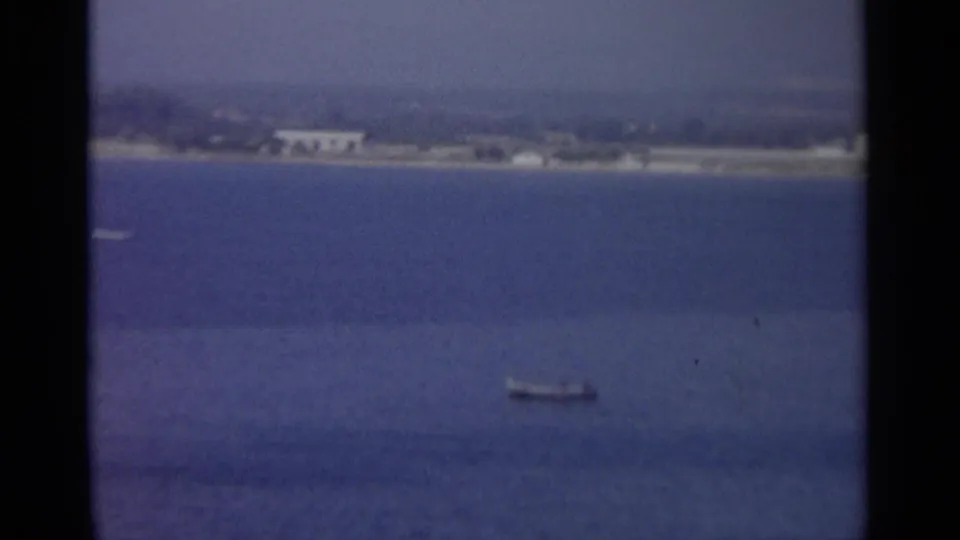 A grainy 8mm view looks across calm blue water toward a distant coast, with a small boat low in the frame.