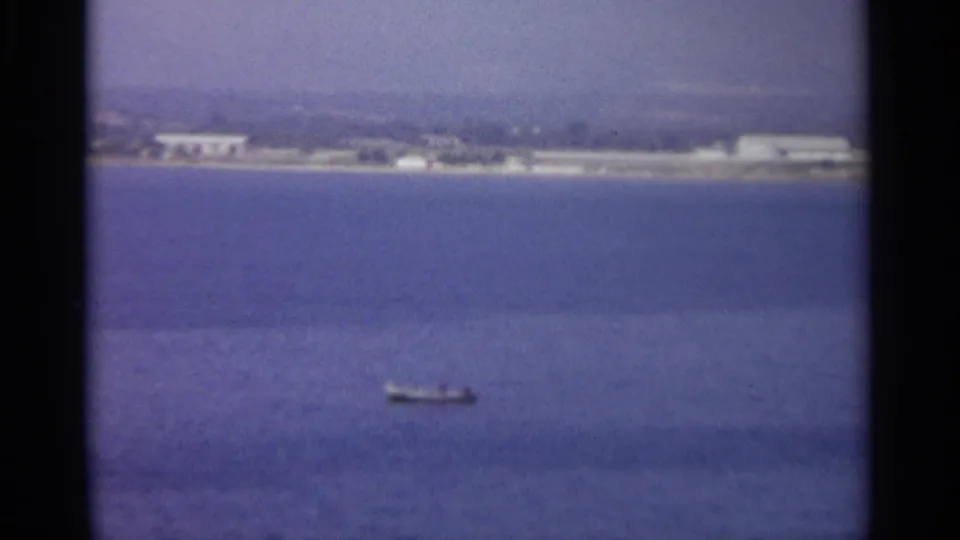 The coastal view continues with a small boat on open water and pale buildings along a hazy Sicilian shoreline.