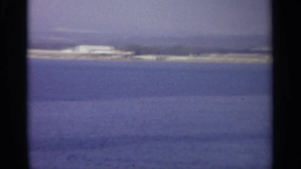 1965 Sicily 8mm frame of a far harbor shore under haze, with blue water filling most of the view.