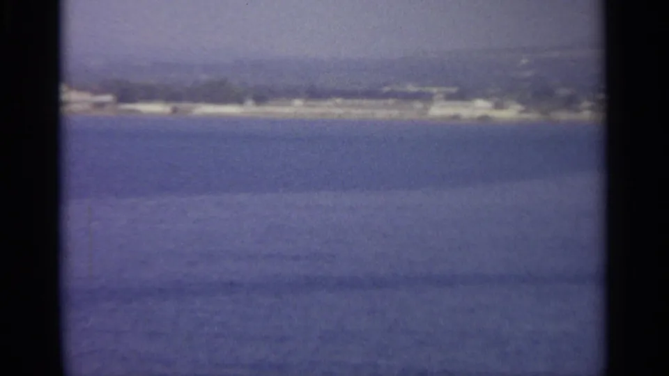 1965 Sicily 8mm frame showing a softened view of water, distant shore buildings, trees, and hazy land beyond.