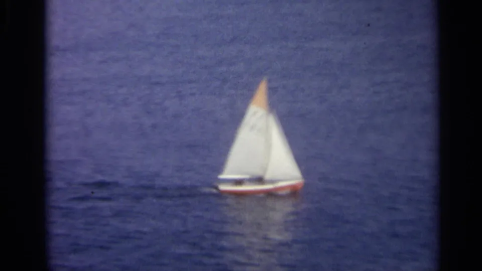 A small white sailboat with an orange sail tip moves across blue water in archival 8mm footage.