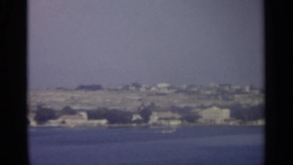 A distant shoreline with pale buildings and low hills appears beyond the water in the home-movie view.