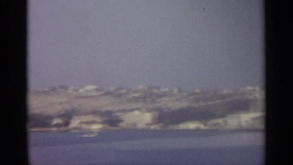 The frame holds on a soft, grainy view of a coastal hillside with scattered pale buildings above the water.