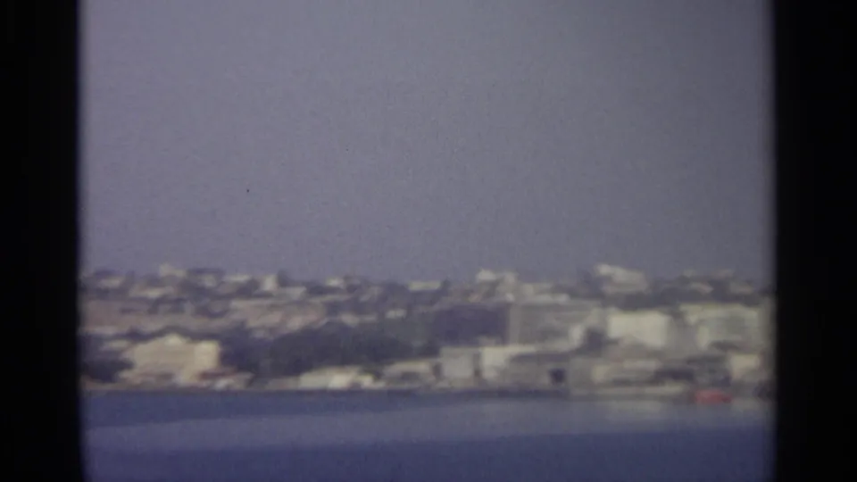 1965 8mm view of a hazy Sicilian coastal city seen across calm blue water, with pale buildings along the shore.