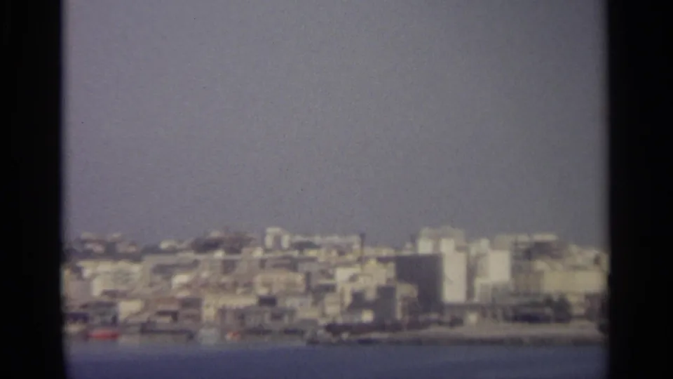 Soft 8mm frame of a Sicilian seaside skyline, with light-colored buildings clustered above the blue water.