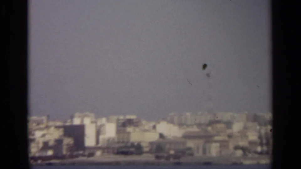 Hazy 1965 8mm view of coastal buildings across the water, with visible film specks and dark contact-sheet borders.