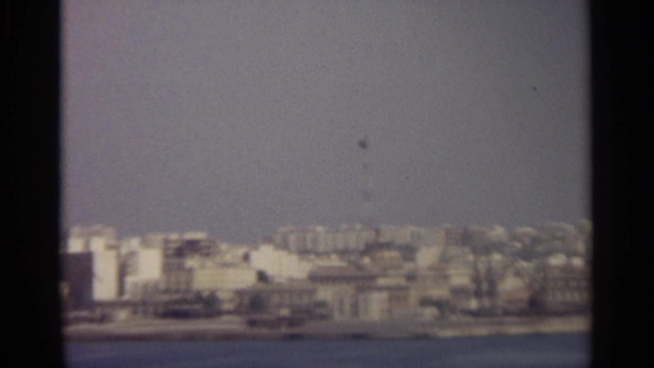 Soft 8mm view of a dense Sicilian waterfront city under a pale blue sky, seen from across the water.